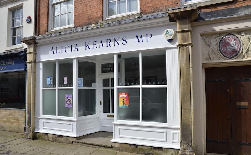 A shop front on a high street with big windows and white-painted panes. A white sign over the entrance reads "Alicia Kearns MP"